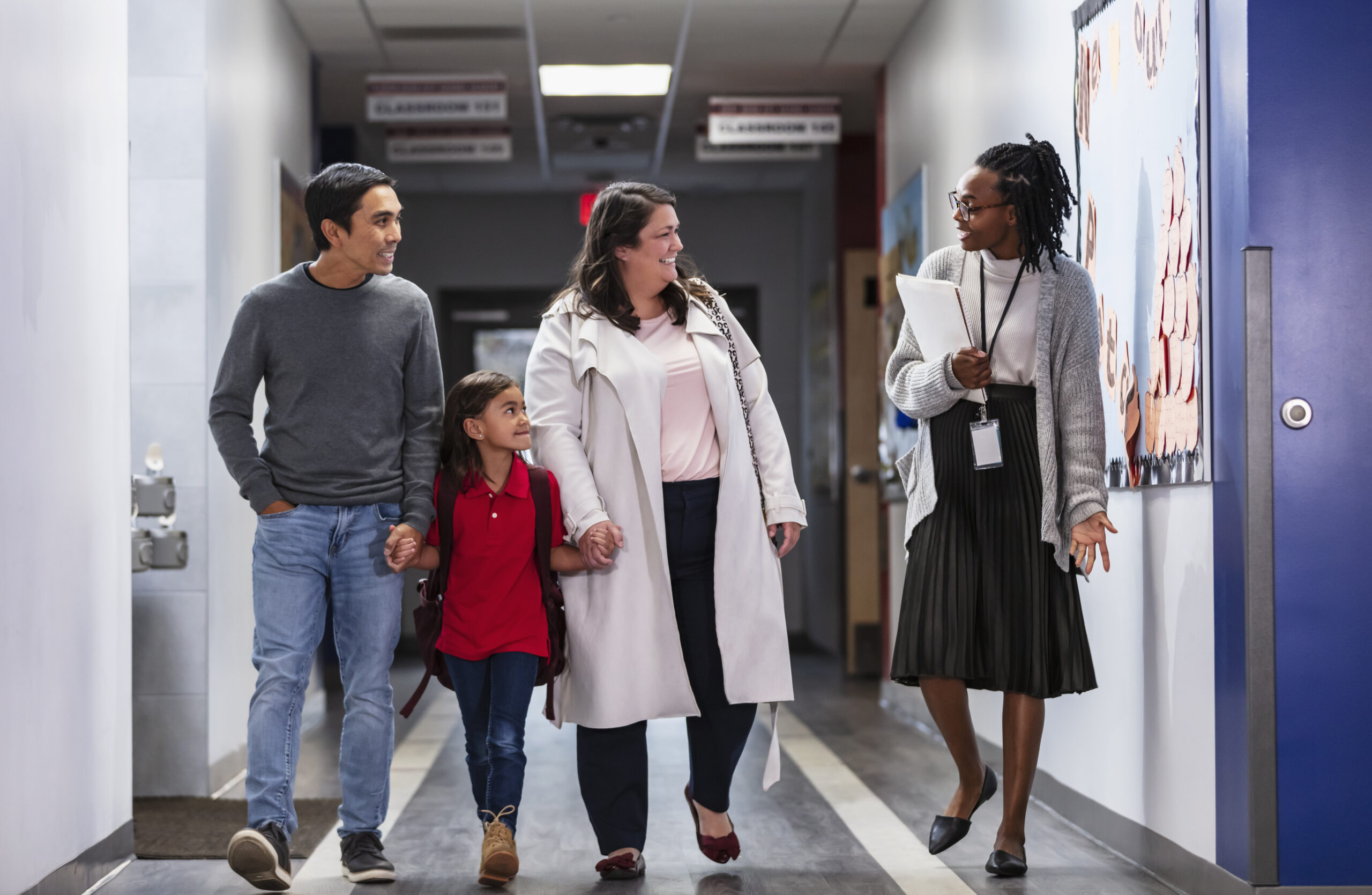 Two parents bringing their daughter to school, talking with an elementary school teacher or principal in the hallway.